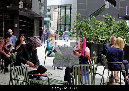 Touristen, die in der City arbeiten aus den unterschiedlichsten Hintergründen in den Stühlen sitzen und essen, Kontakte knüpfen und an Karten Broadgate Kreis, London, UK. Landschaft. Stockfoto