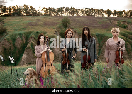 Weiblich musikalischer Quartett mit drei Violinen und Cello bereitet zur Zeit der Blüte Wiese gegen den Hintergrund der malerischen Landschaft neben dem Hund zu spielen. Stockfoto