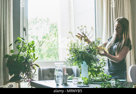 Schöne Frau mit langen blonden Haaren, die die wilden Blumen Strauß in der Vase auf dem Tisch im Wohnzimmer am Fenster. Home Lifestyle und Dekor Ideen Stockfoto