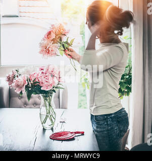 Formschöne Frauen arrangieren Pfingstrosen Bündel in Glas Vase auf dem Tisch am Fenster mit Sonnenuntergang Licht im Wohnzimmer. Lifestyle. Happy Home Stockfoto