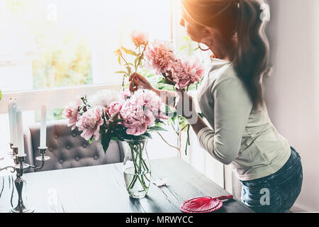 Attraktive Frauen arrangieren Pfingstrosen Bündel in Glas Vase auf dem Tisch am Fenster im Wohnzimmer. Lifestyle. Happy Home Stockfoto