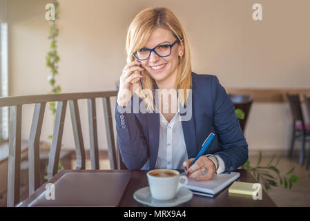 Attraktive Frau mit Brille sprechen auf dem Smartphone, Notizen in einem Café, Restaurant. Charmante Happy girl ist die Vorbereitung für die Arbeit. Auf der Suche nach Stockfoto