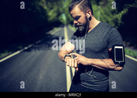 Passen junger Mann in Sportswear steht in der Mitte eines Landes Straße seinen Sport Kontrolle beobachten, bevor sie sich für einen Lauf Stockfoto