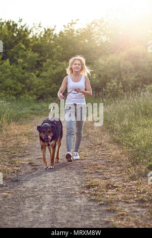 Gerne Frau mittleren Alters zu ihrem Hund entlang einer Grasbewachsenen ländlichen Track im Frühjahr mit Blick auf die Kamera Stockfoto