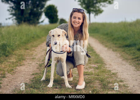 Glückliche junge blonde Frau hocken auf einem ländlichen Dirt Road mit ihren Armen um Ihren Hund und ein glückliches Lächeln Stockfoto