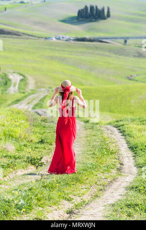 San Quirico d'Orcia, Val d'Orcia, Siena, Toskana, Italien. Eine junge Frau im roten Kleid auf einem Feldweg in der Nähe der Zypressen von Val d'Orcia Stockfoto