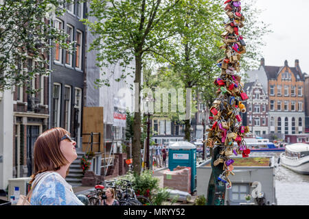 Frau (Alter 40-50) sieht in der Kette der Vorhängeschlösser auf einer Brücke in Amsterdam links Liebe zu gedenken. Kanal und Stadthäusern im Hintergrund. Stockfoto