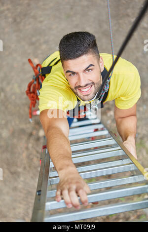 Lächelnden jungen Mann Klettert eine Leiter hinauf in den Hochseilgarten Stockfoto