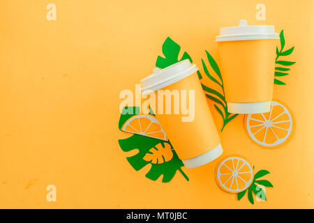 Verfügbare Pappbecher auf gelbem Hintergrund mit tropischen Blättern und Orangenscheiben auf einem gelben Hintergrund. Farbenfrohe Sommer trinken Konzept mit Papier Ausschnitte. Stockfoto