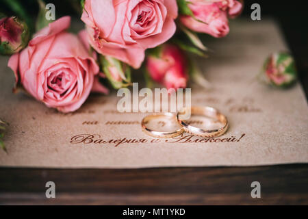 Ein paar goldene Hochzeit Ringe auf der Hochzeit Einladungskarte mit einigen Rosen auf dem Hintergrund. Stockfoto