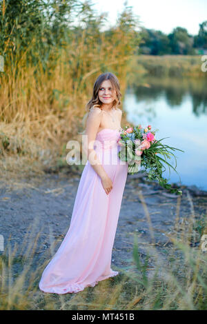 Schwangere junge Frau tragen rosa Kleid mit Blumenstrauß in der Nähe von See. Stockfoto