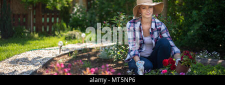 Schöne weibliche Gärtner an Kamera suchen und lächelnd Holzkiste voller Blumen in Ihrem Garten gepflanzt werden. Gartenarbeit Konzept. Stockfoto