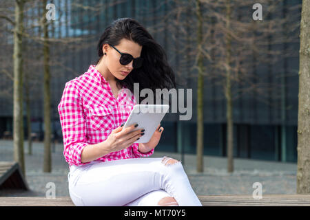 Schöne und konzentrierte sich Mädchen schaut bei der Tablette. Eine Frau in einem rot kariertem Hemd sitzt auf einer Bank im Freien und surft im Internet. Girl shopping Stockfoto