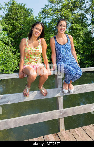 Zwei junge Frauen sitzen zusammen auf der Brücke Geländer oben Teich Stockfoto