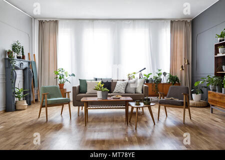 Holztisch mit Blumen zwischen Sessel im geräumigen Wohnzimmer mit Sofa. Real Photo Stockfoto