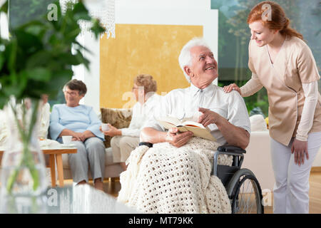 Happy gelähmt älterer Mann im Rollstuhl, ein Buch zu lesen und eine Krankenschwester helfen ihm Stockfoto