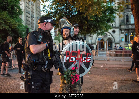 Ein Mann in der bereitschaftspolizei Gang gekleidet mit einem Schild mit 'Frieden' Logo und einem glitzernden Helm, mit einem Blumenstrauß aus Rosen, wirft mit bewaffneten Polizisten nach dem Manchester zusammen Chor Konzert im Gedenken an die Opfer der Arena Bombenattentat in Manchester, Großbritannien, am 22. Mai 2018. Stockfoto