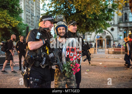 Ein Mann in der bereitschaftspolizei Gang gekleidet mit einem Schild mit 'Frieden' Logo und einem glitzernden Helm, mit einem Blumenstrauß aus Rosen, wirft mit bewaffneten Polizisten nach dem Manchester zusammen Chor Konzert im Gedenken an die Opfer der Arena Bombenattentat in Manchester, Großbritannien, am 22. Mai 2018. Stockfoto