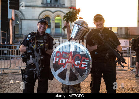 Ein Mann in der bereitschaftspolizei Gang gekleidet mit einem Schild mit 'Frieden' Logo und einem glitzernden Helm, mit einem Blumenstrauß aus Rosen, wirft mit bewaffneten Polizisten nach dem Manchester zusammen Chor Konzert im Gedenken an die Opfer der Arena Bombenattentat in Manchester, Großbritannien, am 22. Mai 2018. Stockfoto