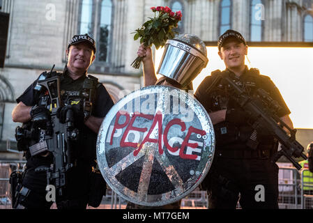 Ein Mann in der bereitschaftspolizei Gang gekleidet mit einem Schild mit 'Frieden' Logo und einem glitzernden Helm, mit einem Blumenstrauß aus Rosen, wirft mit bewaffneten Polizisten nach dem Manchester zusammen Chor Konzert im Gedenken an die Opfer der Arena Bombenattentat in Manchester, Großbritannien, am 22. Mai 2018. Stockfoto