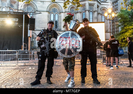 Ein Mann in der bereitschaftspolizei Gang gekleidet mit einem Schild mit 'Frieden' Logo und einem glitzernden Helm, mit einem Blumenstrauß aus Rosen, wirft mit bewaffneten Polizisten nach dem Manchester zusammen Chor Konzert im Gedenken an die Opfer der Arena Bombenattentat in Manchester, Großbritannien, am 22. Mai 2018. Stockfoto