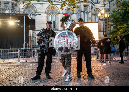 Ein Mann in der bereitschaftspolizei Gang gekleidet mit einem Schild mit 'Frieden' Logo und einem glitzernden Helm, mit einem Blumenstrauß aus Rosen, wirft mit bewaffneten Polizisten nach dem Manchester zusammen Chor Konzert im Gedenken an die Opfer der Arena Bombenattentat in Manchester, Großbritannien, am 22. Mai 2018. Stockfoto