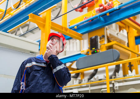 Arbeiter in einer Fabrik arbeiten trägt blaue Anzug und roter Helm suchen in Maschinen im Hintergrund Stockfoto