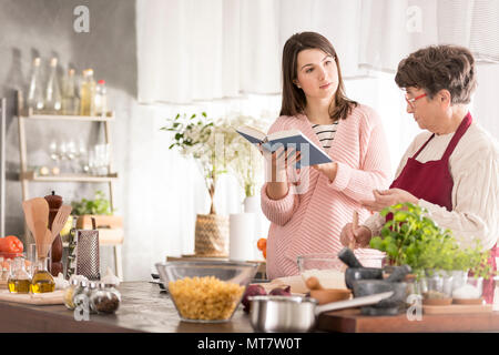 Junge Frau mit einem Kochbuch in einer Küche und im Gespräch mit Oma Stockfoto