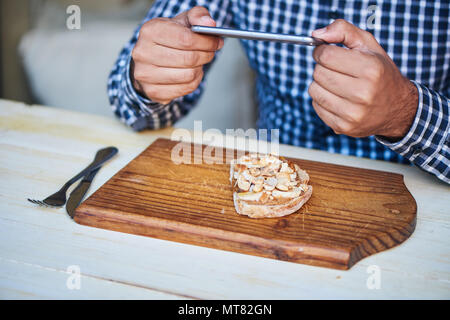 Junger Mann sitzt an einem Bistrotisch mit seinem Smartphone ein Bild von seinem leckeren Erdnussbutter und Banane zu nehmen öffnen konfrontiert Sandwich Stockfoto