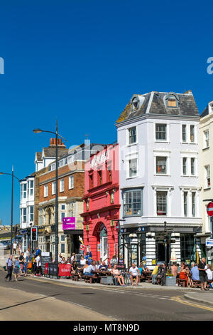 Die Leute draußen sitzen Trinken im Freien auf einer Strandpromenade mit Blick auf die Straße Häuser (die Parade) und das alte Kent Markt Gebäude (rot) in Ramsgate, Kent, Großbritannien Stockfoto