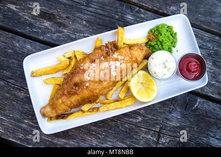 Platte von Panierten Fisch und Chips mit Erbsenpüree und Sauce Tartar, serviert auf einem Holztisch Stockfoto