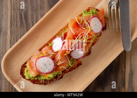 Rustikales Brot Toast mit Guacamole, geräucherter Lachs, Rettich, Brokkoli und Rosenkohl in eine hölzerne Platte auf einem rustikalen Holztisch serviert. Ansicht von oben Stockfoto
