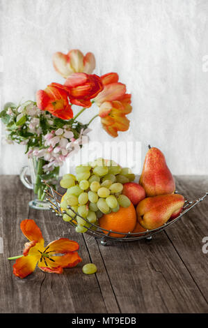 Frucht, Birne, Grapefruit, Zitrone, Pfirsich, Erdbeere in einem Korb auf dem Hintergrund der einen Strauß Tulpen an einem sonnigen Tag. Stockfoto