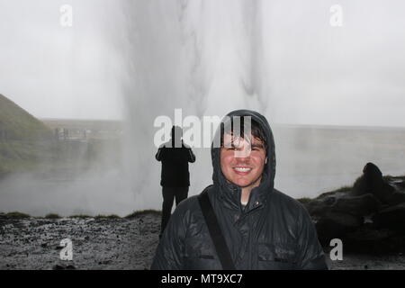 Männliche Touristen stellt sich hinter einem Wasserfall in einen sehr nassen Szene in Island, Island ist für seine eindrucksvolle Landschaft und Möglichkeiten bekannt, mit der Natur in Kontakt zu treten. Stockfoto