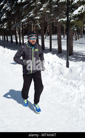 UFA, Russland, 29. MÄRZ 2018 - Junge Mann mit Fahrwerk joggt durch einen Winter Park im Schnee als Teil seiner Winter Sport Fitness Routine fällt. Stockfoto
