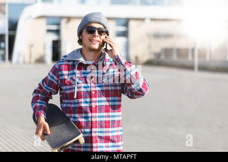Portrait von hipster Mann mit Sonnenbrille im cheked Shirt und Cap holding Skateboard gekleidet und Reden von Smartphone vor modernes Gebäude Stockfoto