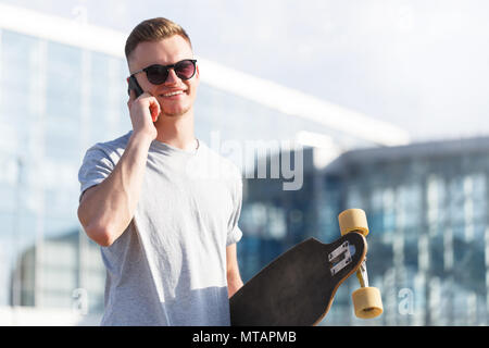 Schön lächelnden jungen Mann im grauen T-Shirt gekleidet und Sonnenbrille holding Longboard und sprechen von Smartphone vor modernes Gebäude Stockfoto