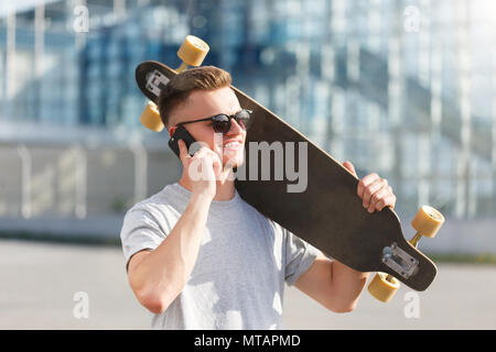 Skateboarder gekleidet in grauen T-Shirt und Sonnenbrille holding Longboard und sprechen von Smartphone vor modernes Gebäude Stockfoto
