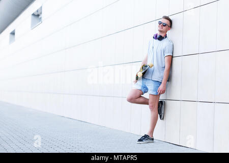 Skateboarder gekleidet in grauen T-Shirt und Sonnenbrille hält Longboard vor modernes Gebäude Ziegel Wand Stockfoto