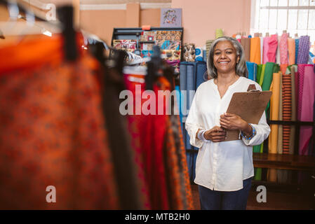 Porträt eines lächelnden reife Stoff shop besitzer neben den Regalen mit bunten Tüchern und Textilien, die eine Zwischenablage Stockfoto