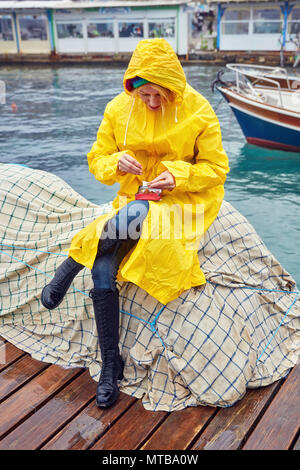 Kaukasische Frau in einem gelben Regenmantel Rollen einer Zigarette auf den Hafen an einem regnerischen Tag. Sie sitzt auf der Netze in der Nähe des Meeres. Stockfoto