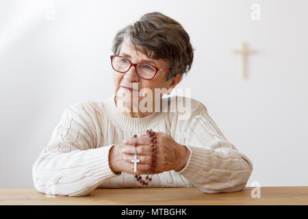 Alte Dame in Gläsern in der Kapelle saß und betete mit Rosenkranz Stockfoto