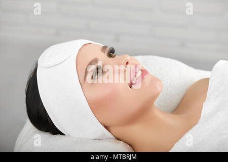 Lächelnde Frau liegend auf weißen cosmetological Trainer tragen headbandage. Warten auf Beaty salon Verfahren und professinal Master. Smoth, junge, gesunde Haut. Wunderschönes Gesicht. Stockfoto