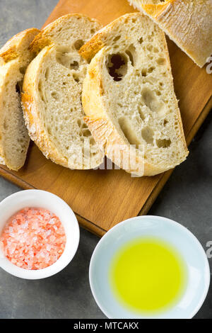 Blick von oben auf die in Scheiben geschnittenen Artisan Rustikales Brot auf Holz Schneidebrett mit Salz und Olivenöl auf dunklem Beton Tabelle. Goldene Kruste Appetitlich schwammig Holey T Stockfoto