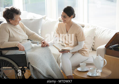 Eine alte Frau im Rollstuhl die Hand eines Angebots Professional Medical Assistant während der Tea Time in einem Wohnzimmer von Luxus in den Ruhestand Stockfoto