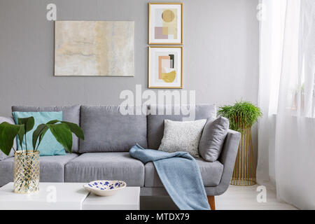 Real Photo von einem grauen Sofa in ein stilvolles Wohnzimmer hinter einem weißen Tisch mit Blätter und vor einer grauen Wand mit Poster Stockfoto