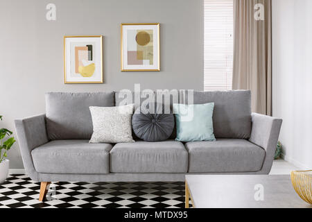 Real Photo von einem grauen Sofa mit schwarzen und weißen Kissen in ein Wohnzimmer Einrichtung mit karierten Boden und Plakate in Gold Frames an einer Wand Stockfoto