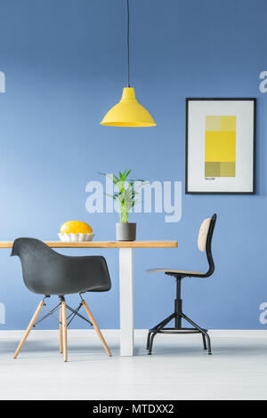Zwei verschiedene schwarze Stühle von hölzernen Tisch mit einer Pflanze und eine Melone in einer Schüssel, eine hängende gelbe Lampe und ein Mockup Poster auf einem blauen Wand in hellen Dini Stockfoto
