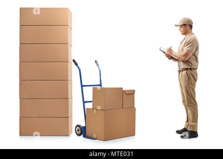 Volle Länge Profil Schuß eines Delivery man neben einem Hand-Lkw und ein Stapel von Kisten schriftlich in einer Zwischenablage auf weißem Hintergrund stehend Stockfoto