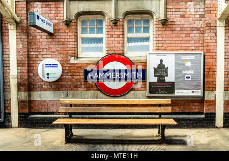 Eine Ikone der Londoner U-Bahnstation name Zeichen für Hammersmith Station auf eine Mauer vor einem leeren Holzbank. Stockfoto